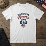White t-shirt with a small stars and stripes heart and the words "My Favorite Veteran Is My Dad", on wood with a flag