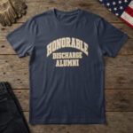 Navy blue t-shirt with cream and brass varsity letters reading "Honorable Discharge Alumni", laid flat on wood with a flag