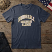 Navy blue t-shirt with cream and brass varsity letters reading "Honorable Discharge Alumni", laid flat on wood with a flag