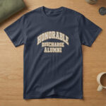Navy blue t-shirt with cream and brass varsity letters reading "Honorable Discharge Alumni", laid flat on wood with a flag