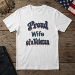 White t-shirt with bold navy and red retro type reading "Proud Wife of a Veteran", laid flat on wood with an American flag