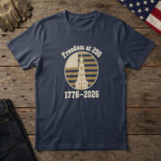 Navy t-shirt with the Independence Hall clock tower on a flag-stripe badge and the words "Freedom at 250 1776-2026", on wood