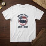 White t-shirt with a solid US map on a flag-stripe badge and the words "We the People 1776-2026", on a colonial desk