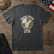 Black t-shirt with a star wax seal and a quill inside a flag badge and the words "Signed, Sealed 1776", flag on wood