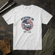 White t-shirt with a navy fire engine on a red and white striped flag badge and the words "STATION 36 EST. 1998"